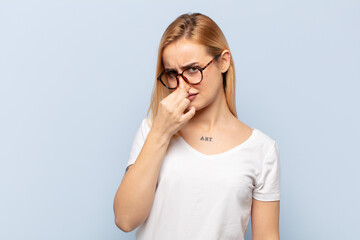 young blonde woman feeling disgusted, holding nose to avoid smelling a foul and unpleasant stench