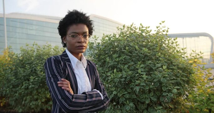 4k. Slow motion. Serious prety Afro-American businesswoman crosses her arms on chest standign before green bushes in the rays of evening sun