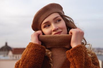 Fashionable happy smiling woman wearing trendy beige beret, turtleneck sweater, brown faux fur coat, posing in European city. Copy, empty space for text
