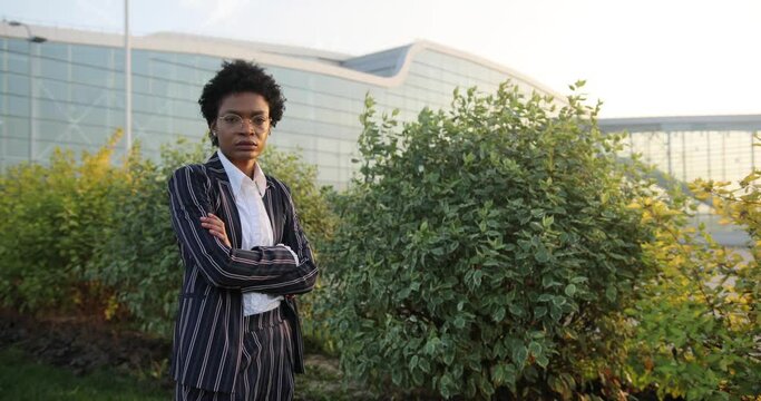 4k. Slow motion. Serious prety Afro-American businesswoman crosses her arms on chest standign before green bushes in the rays of evening sun