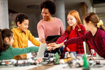 Happy kids with their African American female science teacher with laptop programming electric toys and robots at robotics classroom