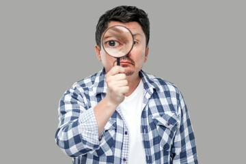 Portrait of serious middle aged man in casual checkered shirt standing, holding magnifying glass and looking at camera with big zoom eye. indoor studio shot, isolated on gray background.