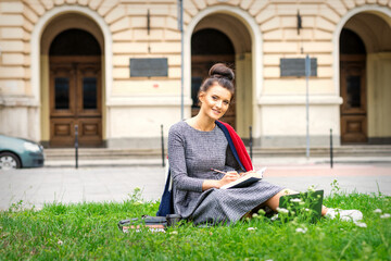 Portrait of a beautiful young smiling student studying with a book on the grass near the university building