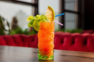 A tiki style orange cocktail in a colorful glass, celery and lemon garnish, at the bar