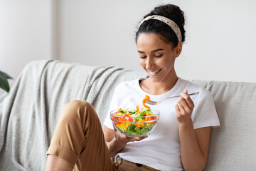 Smiling woman sitting on sofa at home, eating healthy food