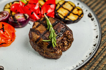 Filet mignon with grilled vegetables and rosemary garnish on a white plate, close-up