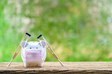 Sick piggy bank wearing surgical mask to prevent viruses and diseases and use underarm crutches put...