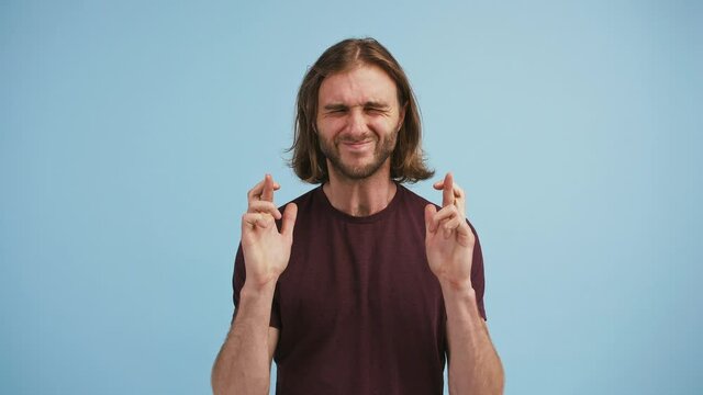 Male folded palms, crossing fingers, looking up and begging please, screaming yes being overjoyed against blue background