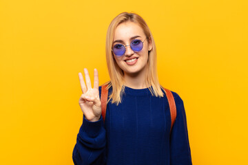 young blonde woman smiling and looking friendly, showing number three or third with hand forward,...