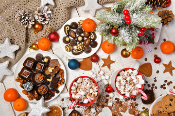 sweet food top view background for merry christmas or new year holiday decoration - chocolate candies, tangerines, cookies, marshmallow and cocoa latte on white wood