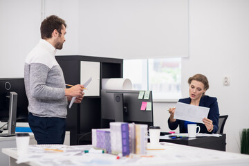 A young male employee receiving directions from his manager. The scene takes place in a bright office, the manager is sitting at her desk and giving instructions refering to some documents, while her