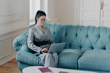 Elegant prosperous female concentrated on online communication, busy doing distance job, sits on comfortable sofa in cozy cabinet, uses modern laptop computer. Journalist writes publication article