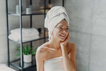 Positive European woman smiles pleasantly, touches healthy skin, wears wrapped towel on head, poses in bathroom. Spa lady with natural makeup poses refreshed after taking shower. Pampering, wellness