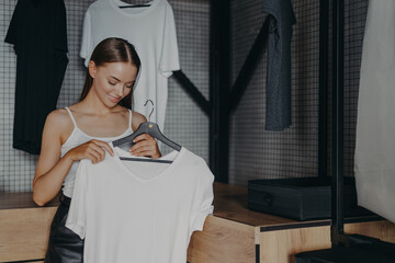 Brunette young woman chooses outfit from home wardrobe, holds white blouse on hangers, picks outfit to wear on specical occasion, poses indoor. Women, dressing, clothing and fashion concept.