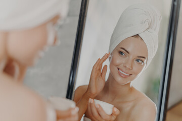 Cute smiling young woman applies beauty cream on face, smiles pleasantly, has minimal makeup, wears wrapped towel on head, looks in mirror, poses in bathroom. Wellness, beauty treatments concept