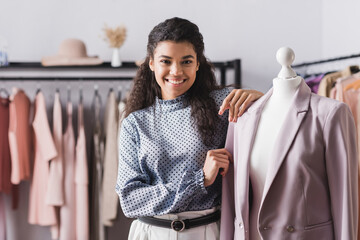 happy african american showroom owner smiling at camera while standing near mannequin with blazer