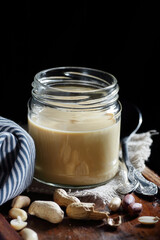 Peanut butter or sauce in a jar on dark moody black background with peanut nuts, spoon and textile nearby, closeup, copy space, healthy vegan food concept, vertical