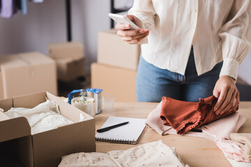 Cropped view of african american owner of showroom using smartphone near box and clothes on table