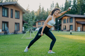 Sideways shot of active smiling determined woman raises dumbbells and has morning workout, dressed in active wear, poses on green lawn against private houses. Sportswoman trains biceps with weights