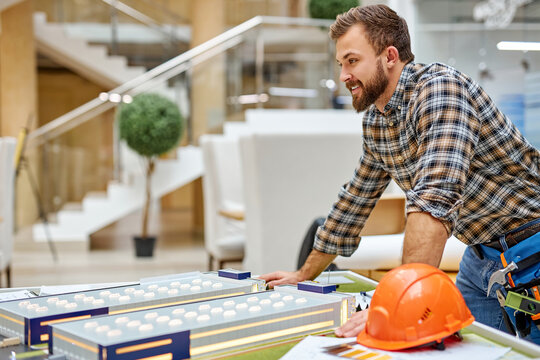 smiling architect enjoy planning the process of building using small 3d model on table, look away , wearing checkered casual shirt, side view