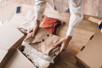 Cropped view of african american businesswoman putting clothes in carton package on table in...