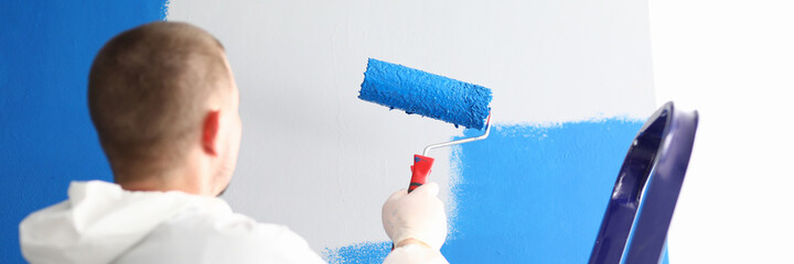 Fototapeta premium Close-up of professional male worker painting wall with blue paint using roller for work. Change interior design in new apartment. Metal ladder. Renovation and construction site concept