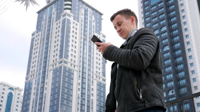 Concentrated handsome man wearing jacket surfs internet in smartphone standing against highrise buildings of residential complex low angle shot