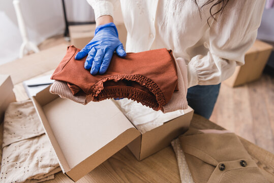 Cropped view of businesswoman in latex glove putting clothes in cardboard box in showroom