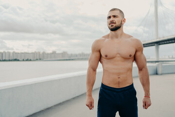 Obraz premium Young bearded athletic man with naked torso muscular body looks somewhere into distance, dressed in sport shorts, rests during workout, poses outdoor near river bridge. Fitness workout concept