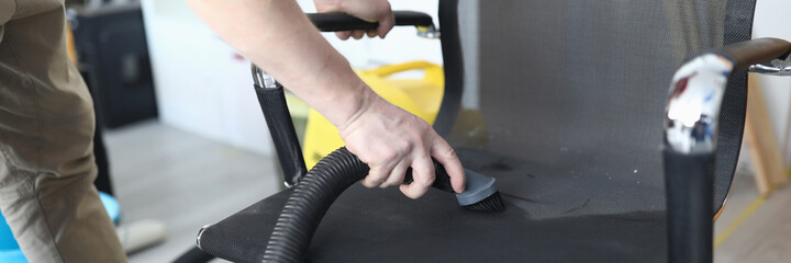 Close-up of person cleaning black comfy chair using compact vacuum cleaner. Place for sitting. Man cleaning in home or office. Electrical brush machine. Cleaning service concept