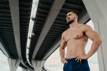 Fototapeta na wymiar Strong man with naked torso keeps hands on waist, has muscular body after long training, focused into distance, stands under bridge, thinks about how to be healthy and fit. Sporty man outdoor