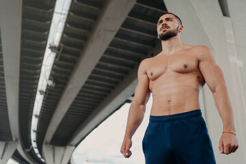 Fototapeta na wymiar View from bottom of self confident bearded handsome European man has strong perfect body looks into distance, leads active lifestyle, poses outside under bridge. Young shirtless bodybuilder.