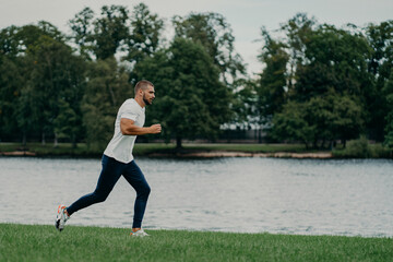 Handsome athlete bearded man runs outdoors in morning poses near river enjoys nature and fresh air, demonstrates endurance and motivation, has morning workout every day, stays fit healthy strong