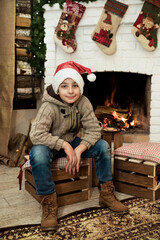 Smiling boy in a knitted sweater and Santa hat on the background of the fireplace and firewood. Christmas mood