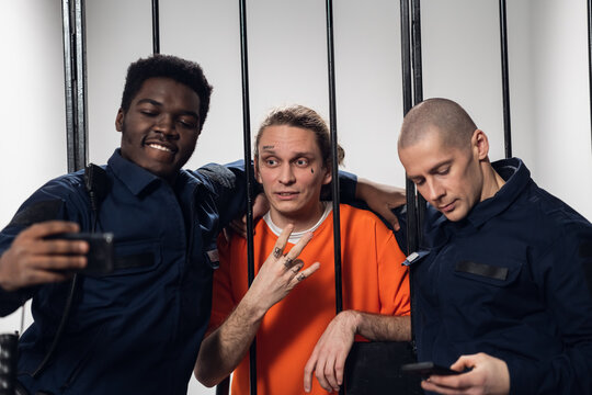 Warders in full gear take selfies on a smartphone with a dangerous criminal behind bars. Black guy and white guy