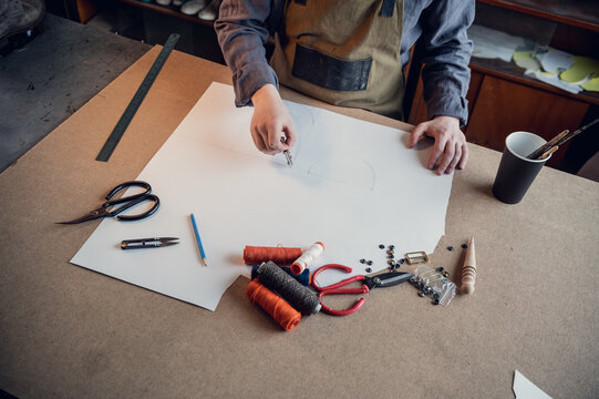 A young shoemaker makes a drawing for a pattern for leather shoes on a table in his workshop.