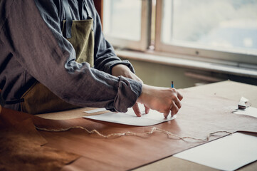 A young apprentice transfers a pattern from paper to leather using a pencil in the workshop on the table.