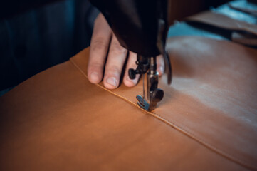 Close-up photo of a high-quality thread seam on a piece of brown genuine leather. Hands of a tailor and a sewing machine.