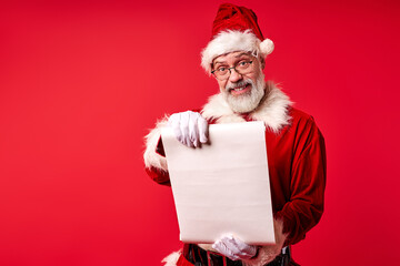 Naklejka premium Santa Claus wearing costume holding parchment roll reading letter wish list, elderly male is preparing for Christmas holiday standing isolated on red background
