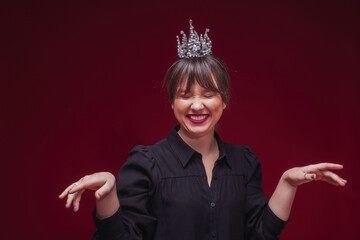 Beautiful laughing model with red lips and closed eyes posing in crown in studio by dark red background  © Lileinaya