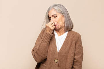 middle age woman feeling disgusted, holding nose to avoid smelling a foul and unpleasant stench