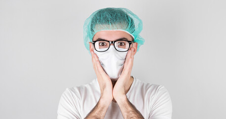Doctor surgeon in white medical mask and a medical cap stands over white background with surprise,holding face with the hands. Сoncept of covid 19, flu and seasonal cold.