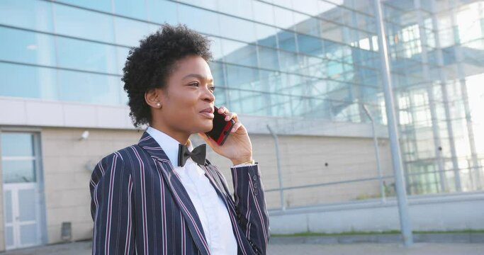4k. Travel, digtal, business. Attractive African American woman in stylish stripped suit talks on the phone walking before a modern glass building