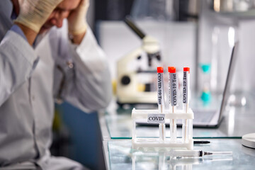 close-up photo of test tubes with coronavirus samples amd scientist male holding head in the background, worried by experiments