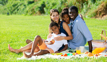 Portrait of cheerful mixed race parents with two children posing together during family picnic on green lawn in summer city park