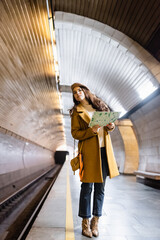 Fototapeta premium stylish woman in autumn clothes looking away while holding city map on underground platform