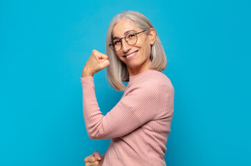 middle age woman feeling happy, satisfied and powerful, flexing fit and muscular biceps, looking...