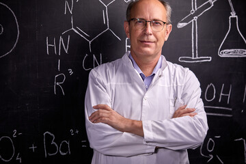 elderly caucasian scientist man posing near blackboard with formulas, looks at camera, in white...