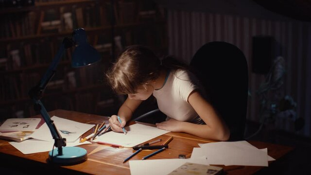 Child schoolgirl draws late evening under light of table lamp at home