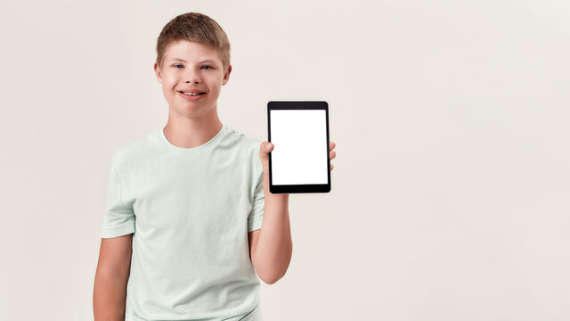 Happy disabled boy with Down syndrome smiling, holding and showing tablet pc with blank screen while standing isolated over white background